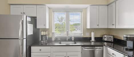 A modern kitchen with white cabinets, stainless steel appliances, a window with blinds, and a black countertop.