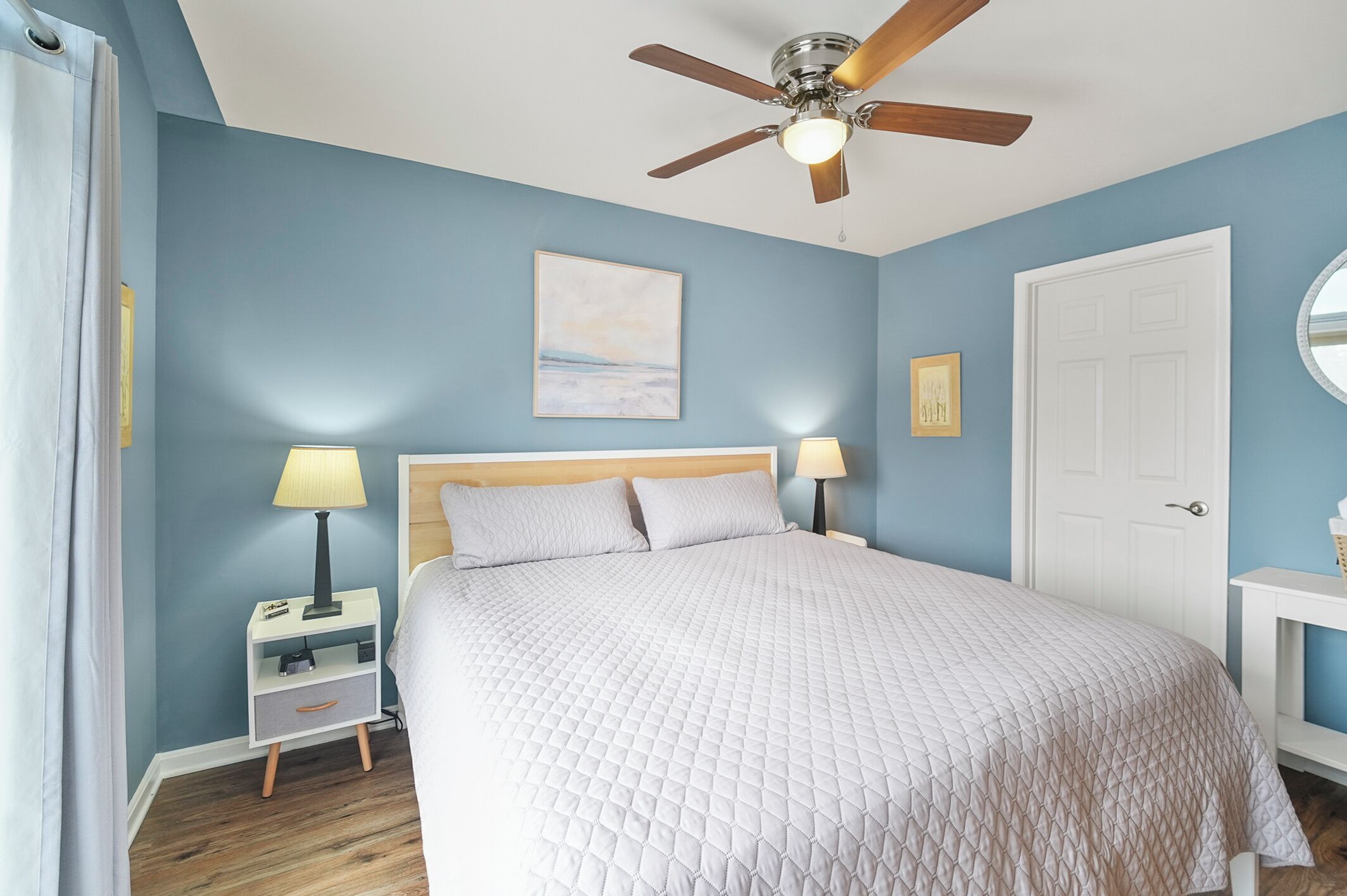 A bedroom with a bed, a nightstand, a lamp, a ceiling fan, a door, and a wall with a painting.