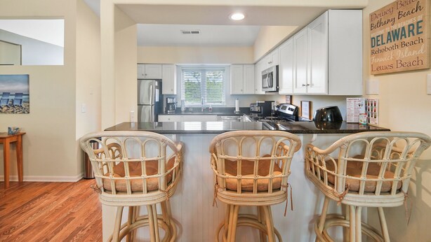 A kitchen with white cabinets, a black countertop, and wicker chairs. - 3BR Lewes Condo Near Beaches | 2 Kings + Screened Porch (Lewes)