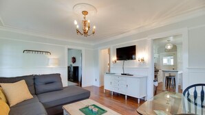 A modern living room with a gray sofa, a glass coffee table, and a dining area with a glass table and chairs. - Hygge Harmony- Salt Hot Tub, 2 Fireplaces, 100+mbps Wifi, AC, Walk to Galveston! (Bend)