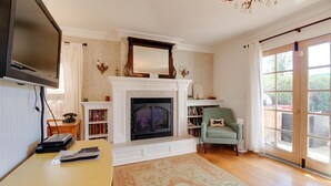 A living room with a fireplace, a television mounted on the wall, a bookshelf, and a large window. - Hygge Harmony- Salt Hot Tub, 2 Fireplaces, 100+mbps Wifi, AC, Walk to Galveston! (Bend)