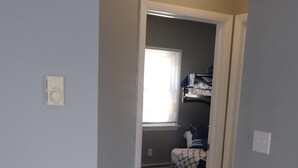 A hallway leading to a bedroom with a patterned rug, a window with blinds, and a bed with a patterned bedspread.
