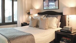 A bedroom with a large bed, wooden beams on the ceiling, a painting on the wall, and a nightstand with books. - Iconic Prospector Condo at Warm Springs (Ketchum)