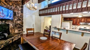 A dining area with a wooden table and chairs, a stone wall, a television mounted on the wall, and a kitchen area visible through an open doorway. - Charming 3-Bedroom, 2-Bath Townhouse at Snow Summit Base – Ski, Hike, Relax! (BIG BEAR LAKE)