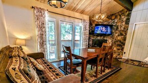 A cozy living room with a wooden ceiling, a stone wall, a television mounted above a fireplace, a dining table with chairs, and a sofa with patterned cushions. - Charming 3-Bedroom, 2-Bath Townhouse at Snow Summit Base – Ski, Hike, Relax! (BIG BEAR LAKE)