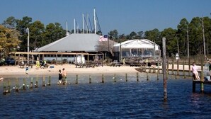 Un bâtiment situé au bord de la plage, doté d’un grand toit et d’une structure adjacente plus petite, qui pourrait être une tente ou un pavillon, le tout entouré d’eau et d’arbres.