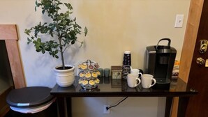 Pieni keittiönurkkaus, jossa on kahvinkeitin, vedenkeitin, ruukkukasvi ja pino mukeja puupöydällä.