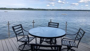 A round table with four chairs on a wooden deck overlooking a calm lake with boats.