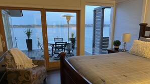 A bedroom with a large bed, a chair, a table, and a view of a body of water through a sliding glass door.