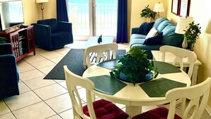 A living room with a dining table, chairs, sofas, a TV, and a view of the ocean through a sliding door. - Fall Steal! 129/nt base! Gulf front, Pool,Huge Balcony,Beach Supplies,VIEW! (Gulf Shores)