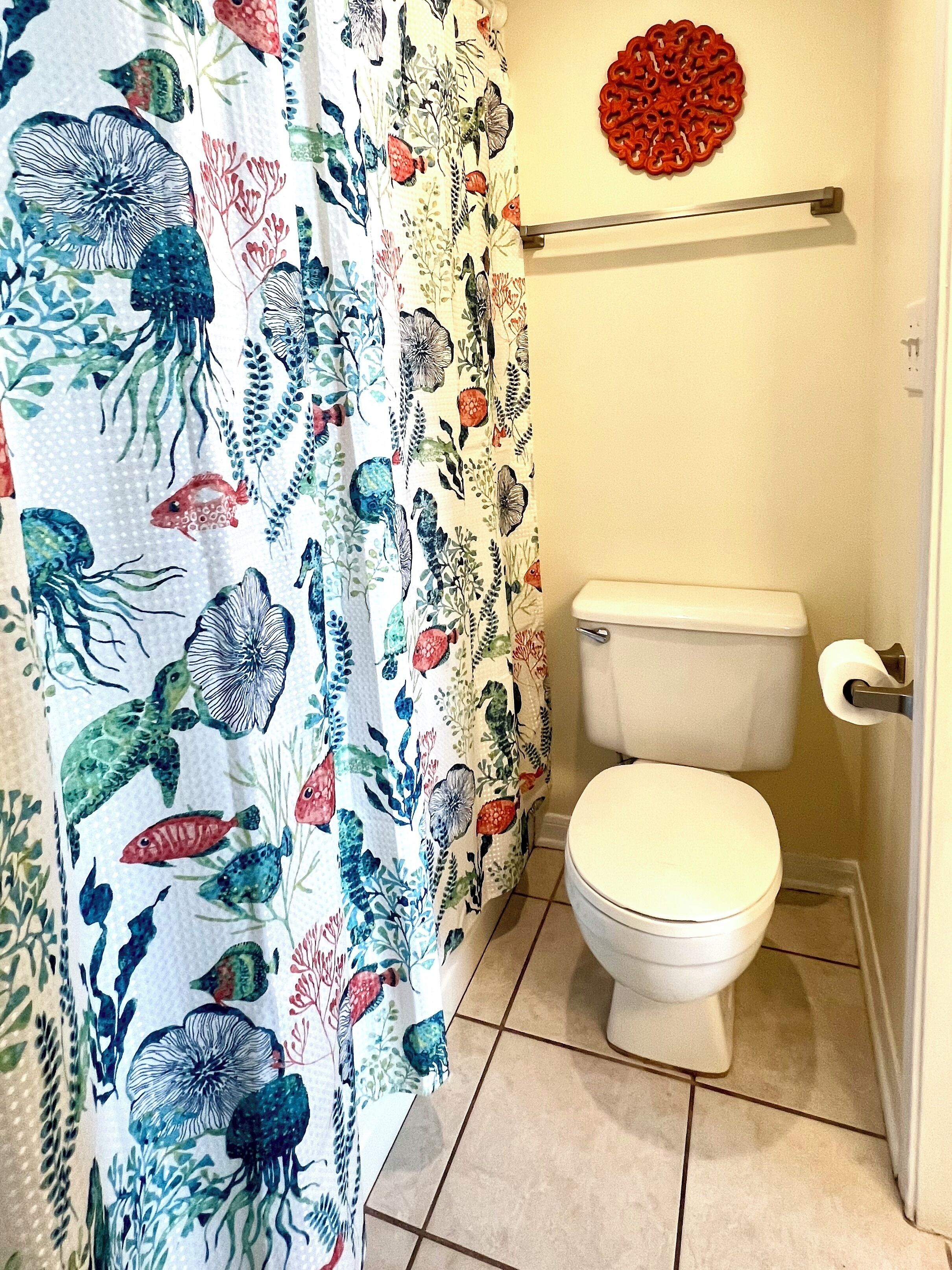 Une salle de bain avec des toilettes blanches, un rideau de douche orné de motifs représentant la vie marine, et un objet décoratif rouge accroché au mur.