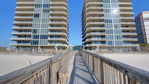A modern beachfront hotel with multiple balconies, a wooden boardwalk leading to the entrance, and a clear blue sky. - Gorgeous Beach Front Corner Unit with Luxury Designer Updates /Mediterranean @PK (Pensacola)