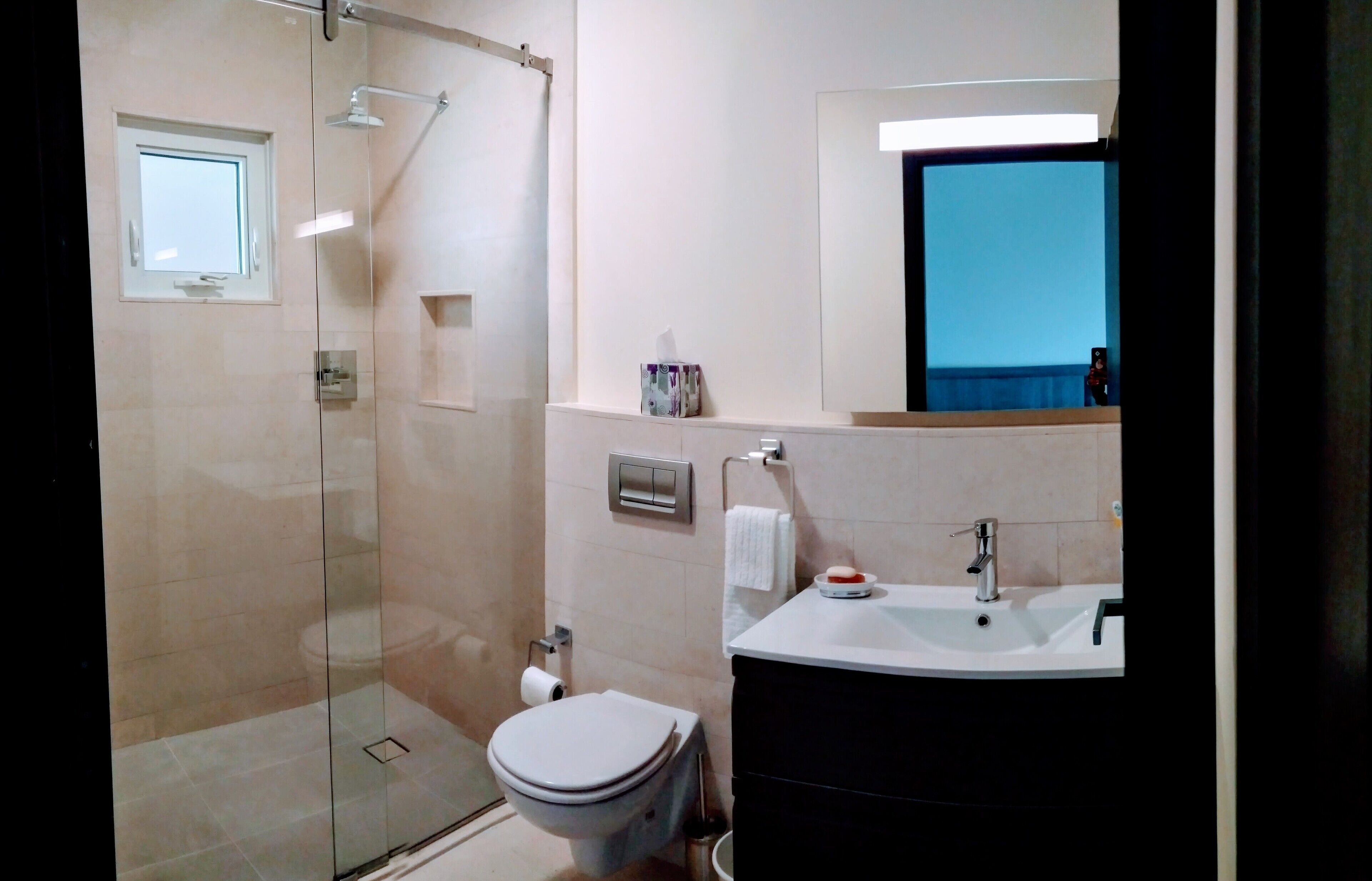 Une salle de bain moderne avec une douche à parois de verre, des toilettes, un lavabo avec un miroir au-dessus et une petite fenêtre.