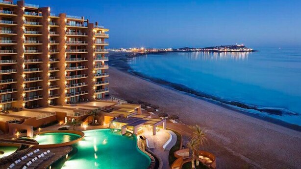 A beachfront hotel with multiple balconies, a swimming pool, and a view of the ocean and distant cityscape. - A View from the Top!  Las Palomas Penthouse Level Beachfront Luxury Condo!!! (Puerto Penasco)