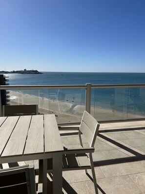 Mesa y banco de madera en un balcón con vista a una playa y una perspectiva despejada del mar y la costa lejana.