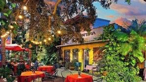 An outdoor dining area with tables and chairs, surrounded by greenery and a building with a sign.