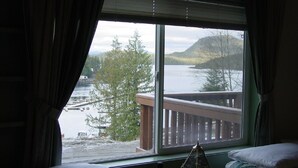 A room with a large window offering a view of a lake, a wooden deck, and a forested hill.