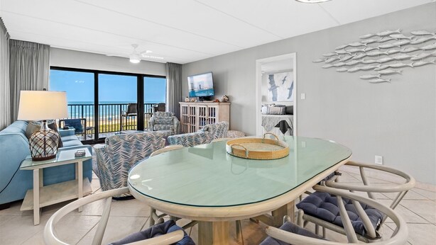 Uma sala de estar moderna com uma mesa de jantar de vidro, assentos confortáveis, uma televisão e vista para a praia.