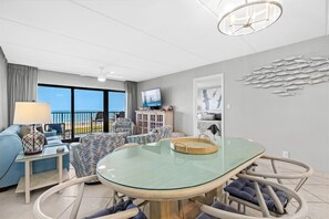 Un salon moderne avec une table à manger au plateau en verre, des sièges confortables, un téléviseur et une vue sur la plage.