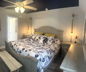 Quarto com cama, mesas de cabeceira, um banco, um ventilador de teto e uma porta.