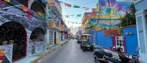 A narrow street lined with colorful buildings, some with murals and decorations. There are parked vehicles and a few people visible.