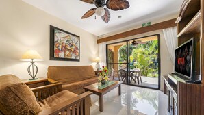 Une chambre d’hôtel avec un canapé, des fauteuils, une table basse, un tableau, une télévision et un balcon donnant sur un paysage verdoyant.