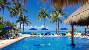 Une piscine entourée de chaises longues et de parasols, bordée de palmiers et surplombée d’un ciel bleu azur.