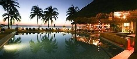 A poolside area with a reflection of palm trees and a thatched-roof structure, likely a bar or lounge, at twilight.