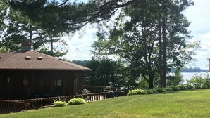 A wooden cabin with a deck, surrounded by a grassy lawn and trees, overlooking a body of water. - Waterfront Lake House on Scenic Lake Nokomis (Tomahawk)