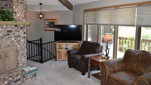 A living room with a brick fireplace, a television, leather sofas, and a large window with blinds. - Waterfront Lake House on Scenic Lake Nokomis (Tomahawk)