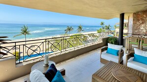 Een balkon met uitzicht op het strand en de oceaan. Er staan twee witte banken met blauwe kussens, een houten tafel en een kandelaar van glas.