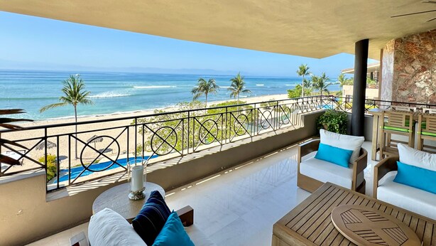 Un balcón con vista a la playa y al océano. Hay dos sofás blancos con cojines azules, una mesa de madera y un candelabro de cristal.