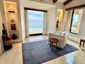 A modern living room with a large window overlooking a beach, a white sofa, a small wooden table, and a decorative shelf with items. - FRONT ROW PENTHOUSE - Amazing views -  Premier/Cook/Golf Cart (Punta De Mita)