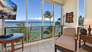 A room with a large window overlooking a beach, a chair, a table, and a TV. -  3 Bedroom, 3 Bath Oceanfront Condo With Amazing Views of Molakai and Lanai  (Lahaina)