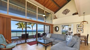 A spacious living area with a large gray sofa, a wicker chair, and a glass coffee table. A balcony with a view of the ocean and palm trees is visible. -  3 Bedroom, 3 Bath Oceanfront Condo With Amazing Views of Molakai and Lanai  (Lahaina)