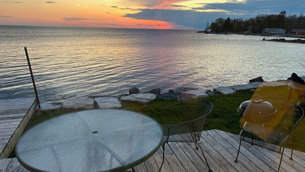 Una terraza de madera con mesa de vidrio y sillas de metal, con vistas a un cuerpo de agua al atardecer.