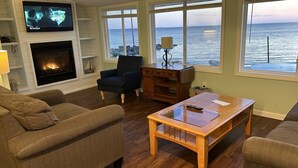 Una sala de estar con chimenea, televisor y ventanas amplias con vista al mar.