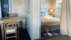 Una habitación con una cama, una mesita de noche con lámpara, una ventana con cortinas, una puerta que da a un baño y una zona de comedor con mesa y sillas.
