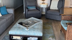 Un salon comprenant un canapé, deux fauteuils, une table basse et une chaise en osier. On y trouve également un ventilateur de plafond, des œuvres d’art murales et, au fond, une cuisine.