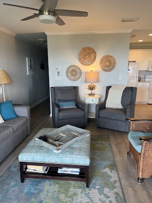 A living room with a sofa, two armchairs, a coffee table, and a wicker chair. There is a ceiling fan, wall art, and a kitchen in the background.
