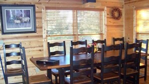 A wooden interior with a dining table surrounded by chairs, a framed picture on the wall, and a window with blinds.