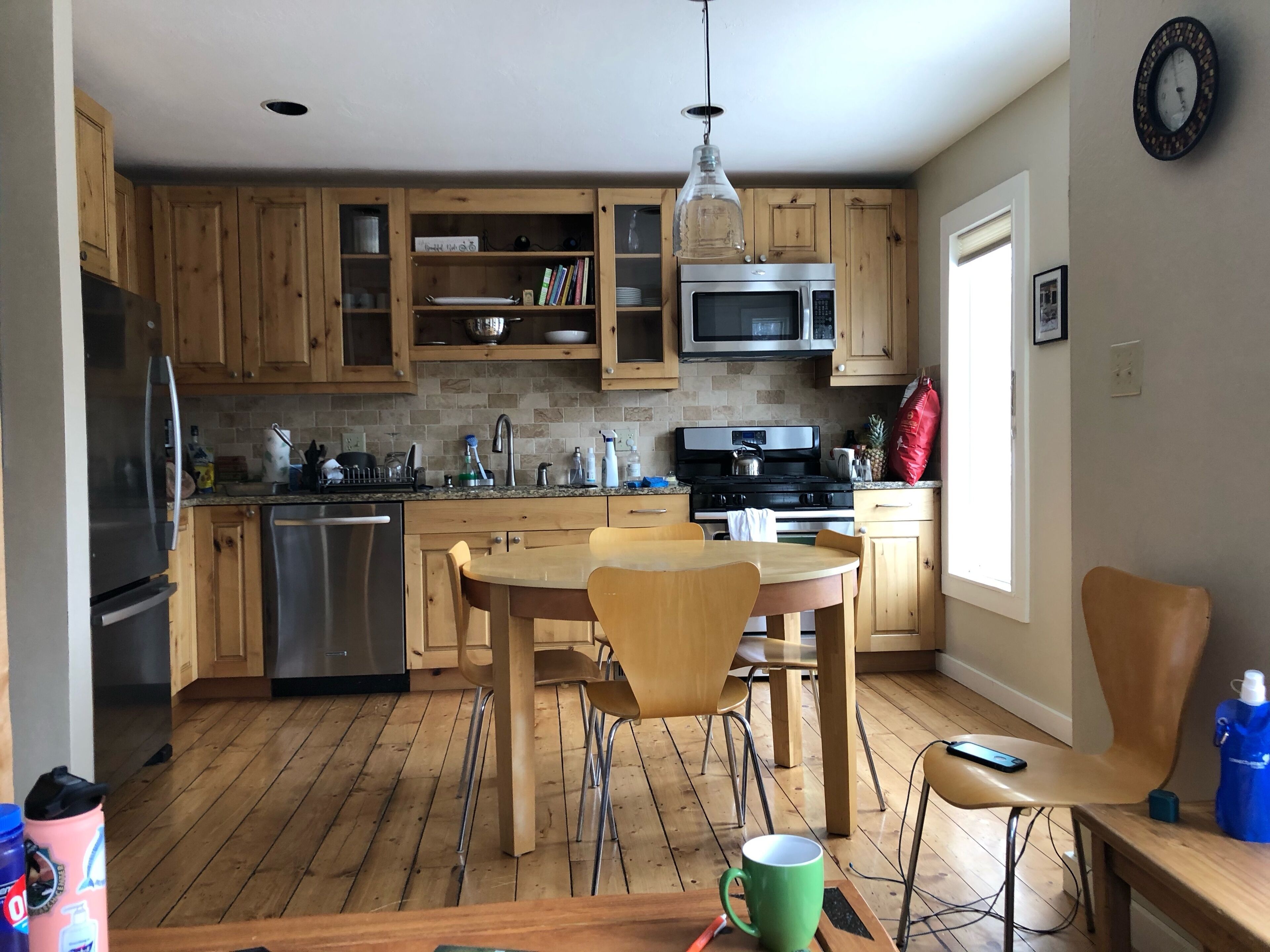 A modern kitchen with wooden cabinets, a stainless steel refrigerator, a microwave, and a stove. There is a dining table with chairs and a hanging light fixture.