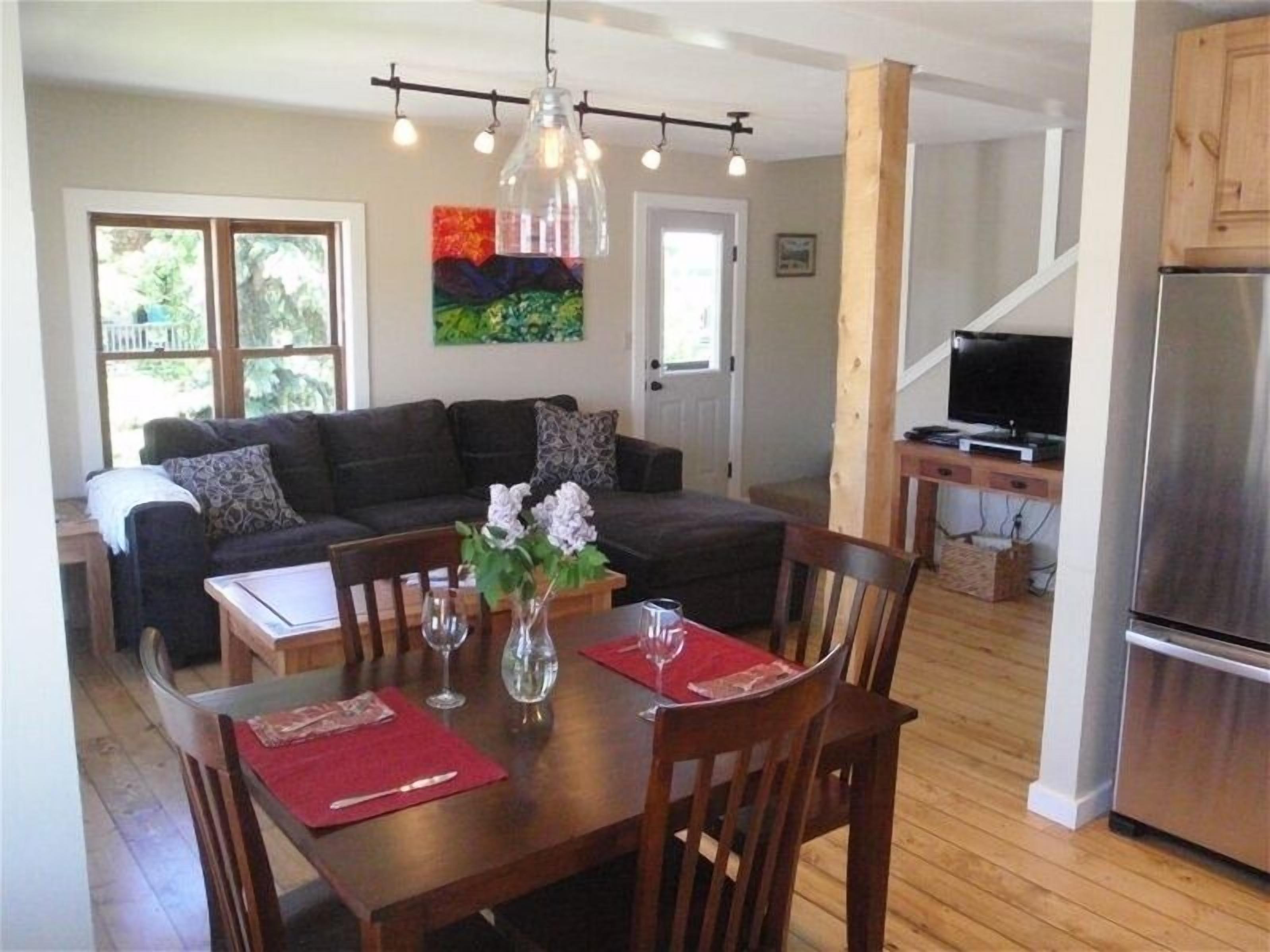 A dining area with a wooden table, chairs, and a vase of flowers. There is a dark-colored sofa in the background and a kitchen area with a refrigerator and a television.