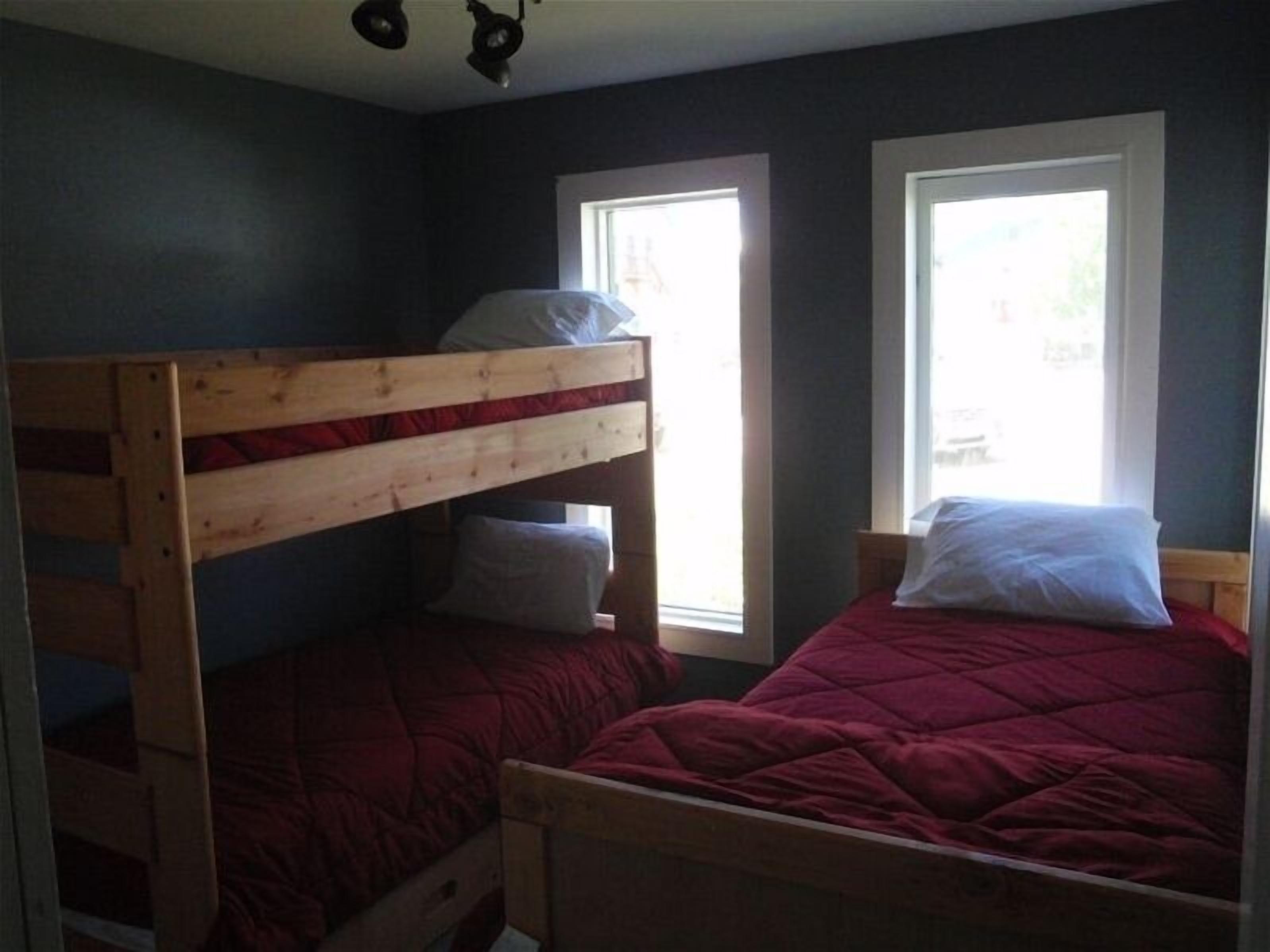A room with two bunk beds, one with a red blanket and the other with a white pillow. There are two windows providing natural light.
