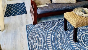 A modern living room with a sofa, a coffee table, and a patterned rug. There is a kitchen area in the background with white cabinets and a dining table.