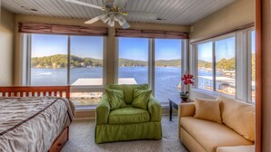 Ein Zimmer mit einem Bett, einem grünen Sessel und einem beigefarbenen Sofa, mit großen Fenstern und Blick auf einen See und Hügel.