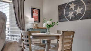 Un comedor con una mesa redonda de madera, cuatro sillas de madera, un jarrón con flores blancas, un cartel enmarcado en la pared y una lámpara sobre una mesita auxiliar.
