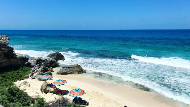 Ein Strand mit klarem, blauem Wasser, Felsformationen und Sonnenschirmen.