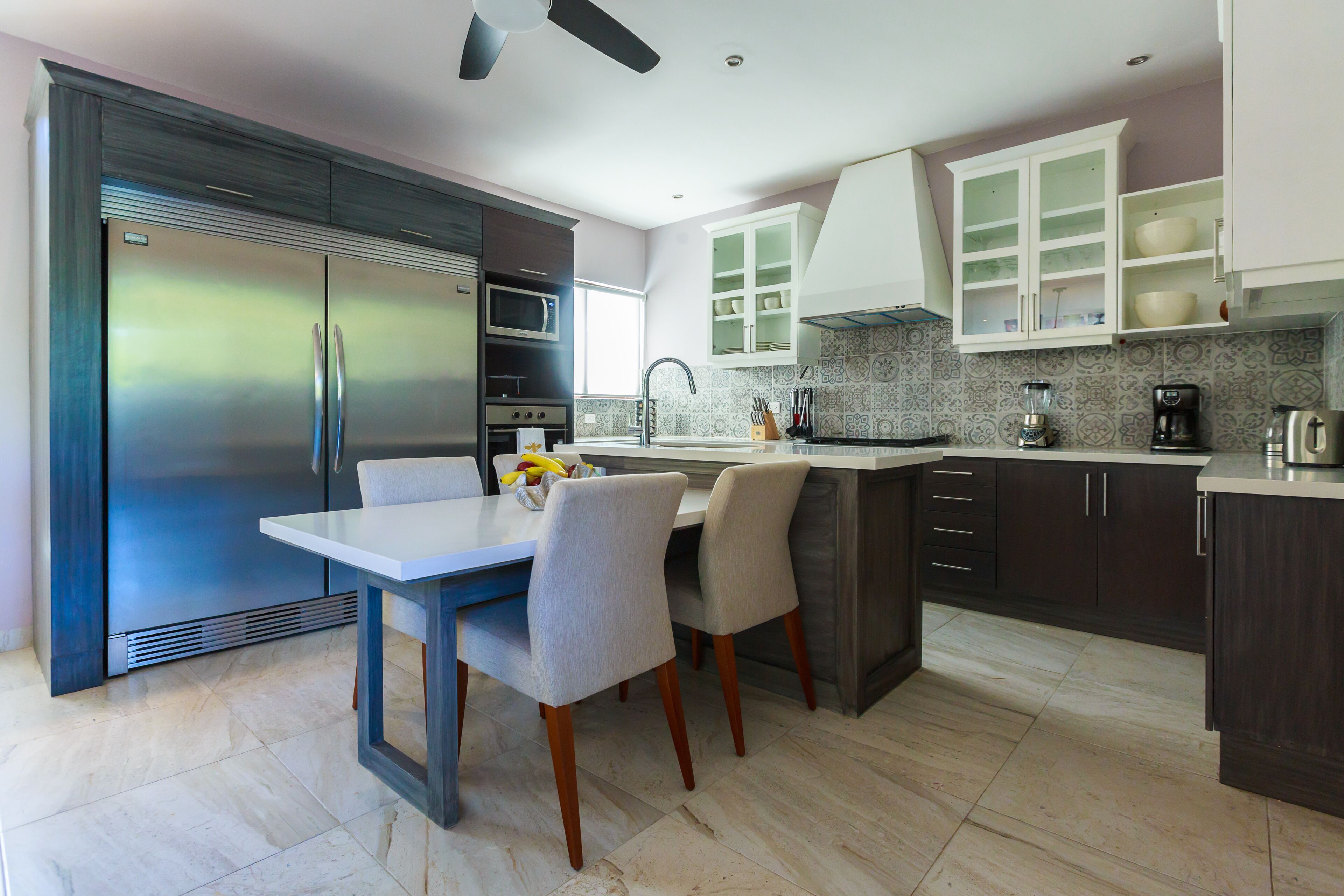 A modern kitchen with a stainless steel refrigerator, a microwave, and a sink. There is a dining table with chairs and a ceiling fan.