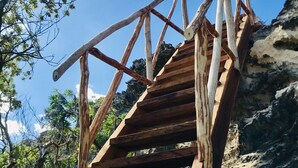 Eine hölzerne Treppe führt über steiniges Gelände, umgeben von Bäumen und unter einem klaren Himmel.